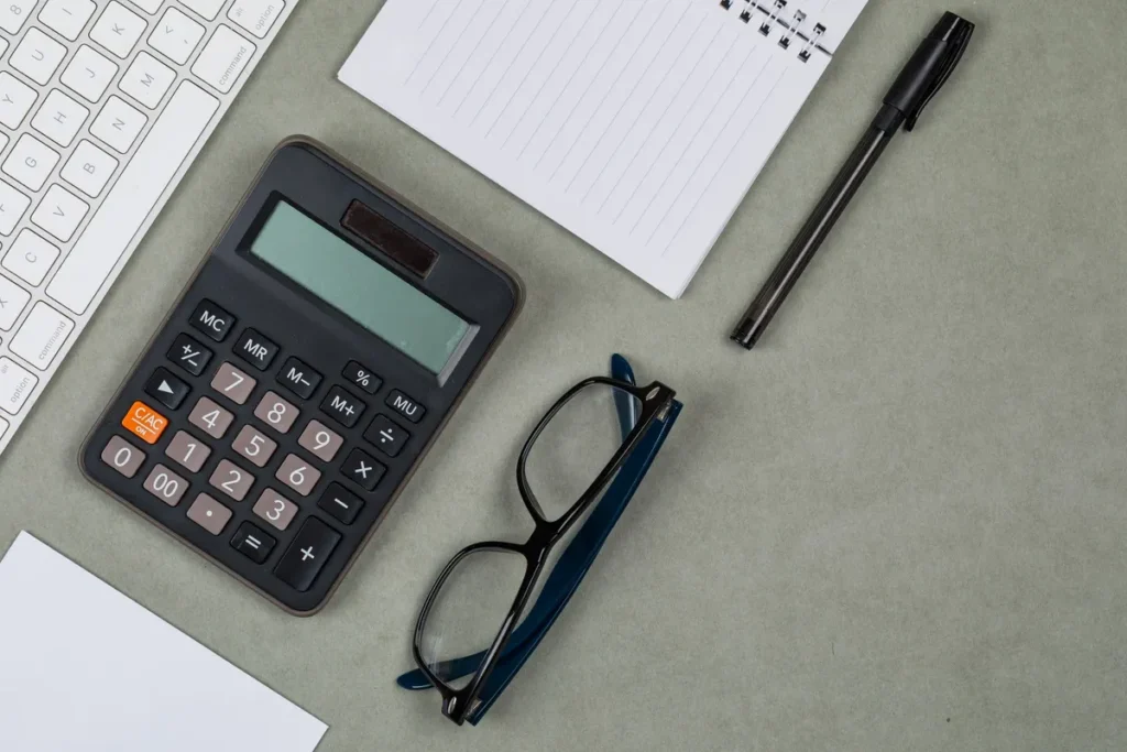 Flat lay of a notebook, pen, calculator, and glasses on a grey background, symbolizing bookkeeping for businesses.